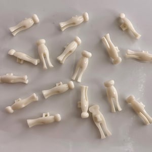 May include: A collection of small, off-white plastic cord locks. Each lock has a cylindrical head, a rectangular body, and two legs. The locks are scattered across a light-colored surface.