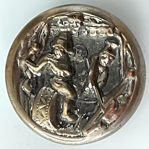 May include: A detailed, antique button with a raised relief design. The button features a scene with multiple figures, possibly depicting a historical or theatrical event. The button has a silver-toned metal finish with areas of gold-toned accents.