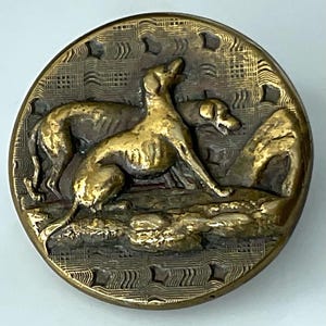 May include: A round, antique brass button featuring a detailed relief of two dogs in a landscape. The dogs are golden in color, with one in a seated position and the other standing. The background has a textured pattern.