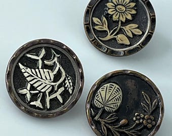 May include: Three antique buttons with floral designs. Each button features a dark, textured background with a raised, light-colored floral motif. The buttons have a metallic, aged appearance, with a decorative edge.