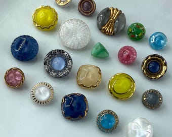 May include: A collection of assorted decorative buttons in various shapes, sizes, and colors, including yellow, blue, pink, green, white, and grey. The buttons feature diverse textures, metallic rims, and smooth or faceted surfaces arranged together.