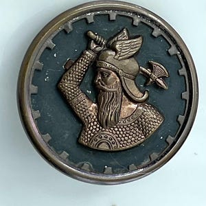 May include: A round, bronze-colored metal button with a Viking warrior design. The warrior is depicted in profile, holding a hammer and sword, with a winged helmet. The background is dark teal, and the button has a gear-like border.