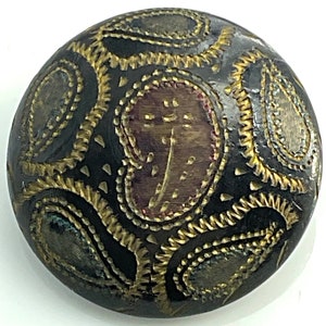 May include: A round, black button with a gold and red paisley design. The button has a raised, textured surface.
