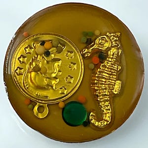 May include: A round, gold-colored button with a seahorse and a coin design. The button has a clear, amber-colored resin background with colorful beads and a green bead. The seahorse and coin are gold-colored.