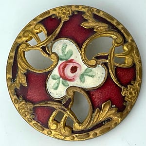 May include: A round metal button featuring an intricate gold-tone filigree frame with deep red enamel fill. The center displays a white triangular inlay decorated with a small hand-painted pink rose and green leaves.
