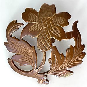 May include: A detailed, bronze-colored floral brooch. The brooch features a large flower with etched details, surrounded by intricately carved leaves and stems. The design is symmetrical and has a vintage aesthetic.