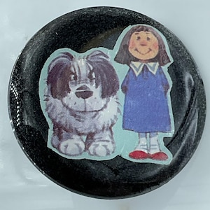 May include: A black, round button with a glossy finish. The button features a cartoon illustration of a girl in a blue dress and a fluffy dog. The girl is wearing red shoes. The dog is black, white, and gray.
