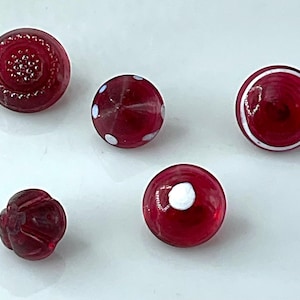 May include: Five small, round, translucent red glass beads. One bead has a textured dotted pattern, one features white polka dots, one has a white circular ring, one is a plain smooth sphere with a white dot, and one has a molded floral shape.