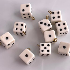 May include: A collection of ivory-colored dice with black dots, each featuring a small gold-colored loop. The dice are scattered, showcasing different number combinations. Ideal for jewelry making or crafting.