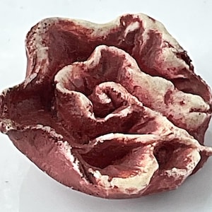 May include: A handcrafted, abstract sculpture of a rose. The sculpture is primarily a deep red color with accents of off-white. The rose is intricately detailed, with visible textures and curves, suggesting a natural, organic form.