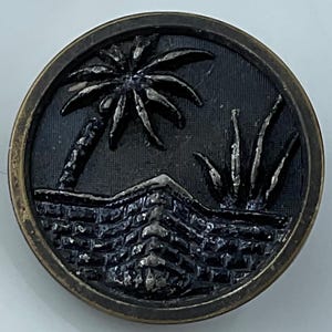 May include: A round, antique-style button with a detailed design. The button features a palm tree, brick wall, and other foliage, all in a dark gray color. The outer rim has a bronze finish, adding to the vintage aesthetic.