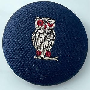 May include: A circular fabric-covered button in a deep navy blue color. The center features an embroidered owl design in white thread with prominent red accents on the eyes and feet, perched on a small branch, creating a detailed contrast.