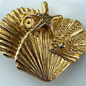 May include: A gold-toned brooch featuring a cluster of three textured seashells and a starfish. The design includes a small circular indentation on one shell and a single, small, smooth silver-colored bead accenting the surface of another shell.