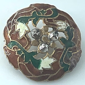 May include: A round metal brooch featuring a brown, green, and white enamel floral pattern with gold-toned outlines. The center of the piece is adorned with three small, faceted clear stones and three smooth gold-toned metal spheres.