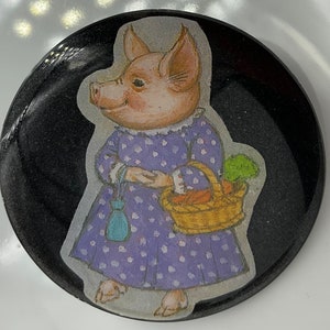 May include: A round button with a black background and a cartoon image of a pig wearing a purple dress with white polka dots. The pig is holding a basket of carrots and a blue purse.