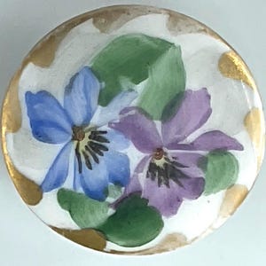 May include: A round, white ceramic button with a hand-painted floral design. The button features two violets, one blue and one purple, with green leaves. The edge is trimmed with gold paint.