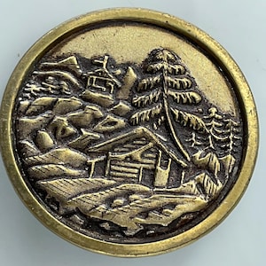 May include: A round, antique brass button with a detailed landscape scene. The design features a cabin, trees, and mountains, all intricately carved. The button has a raised edge and a slightly worn appearance.