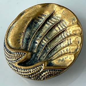 May include: A vintage, oval-shaped button with a textured, metallic gold finish. The design features a stylized feather or wing motif with dark blue accents. The lower edge has a detailed, perforated pattern.