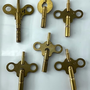 May include: A collection of antique brass clock keys in various shapes and sizes. Some keys have a circular or figure-eight design, while others are more elongated. One key has the number "4" written on it. The keys are arranged on a white surface.