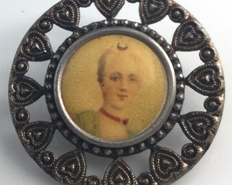 May include: Vintage silver-toned brooch with a central portrait of a person. The portrait is surrounded by a silver frame and a decorative openwork border of hearts. The person in the portrait is wearing a green top and a red necklace.