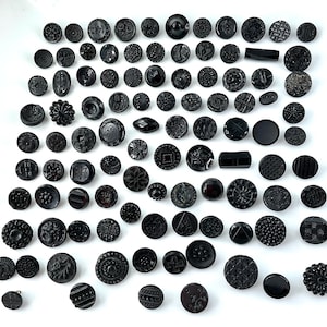 May include: A large collection of assorted black buttons in various shapes, sizes, and intricate patterns. The set includes round, rectangular, and decorative buttons with embossed floral, geometric, and textured designs, all featuring a glossy finish.