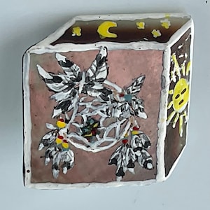 May include: A hand-painted, rectangular decorative tile with a dreamcatcher design. The tile features a pink background with a white dreamcatcher, black and white feathers, and yellow accents. The edges are outlined in white and brown, with a yellow sun and moon.