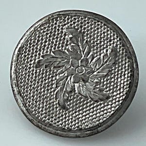 May include: A round, silver-toned button with a textured background and a floral design in the center. The button's surface has a detailed pattern, and the floral element is raised, creating a three-dimensional effect.