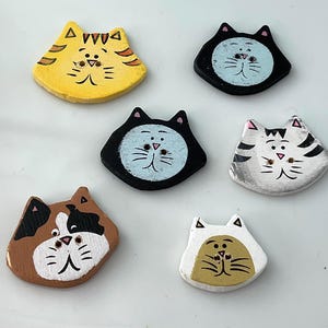 May include: Six flat, cat-shaped buttons with two holes in the center of each face. The buttons feature various colors and patterns, including yellow, black, white, brown, and gray, with hand-painted details like whiskers, noses, and ears.