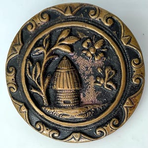 May include: A round button featuring a raised gold-toned relief of a beehive, a tree, and a flying bee set against a recessed black background. The outer border is decorated with a gold-toned scrollwork pattern and triangular accents.
