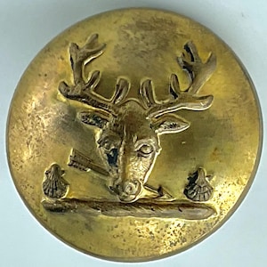 May include: A round, antique brass button featuring a detailed relief of a stag's head with antlers, an arrow, and two scallop shell emblems. The button has a slightly tarnished, aged appearance, suggesting it is vintage.