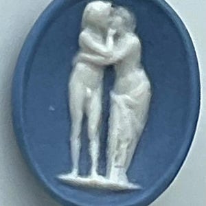 May include: An oval-shaped blue cameo featuring a raised white relief of two figures standing and embracing. The figures are depicted in a classic style with one figure appearing nude and the other draped in fabric, set against a matte blue background.