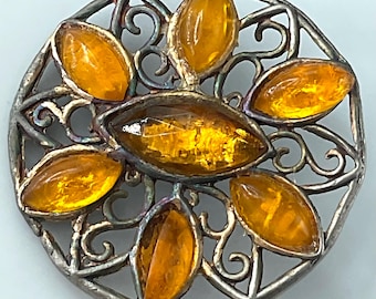 May include: A round, vintage brooch with a silver-toned filigree design. The brooch features seven amber-colored, oval-shaped stones set within the filigree. The stones are arranged in a circular pattern, with one in the center and six surrounding it. The brooch has a classic, antique aesthetic.