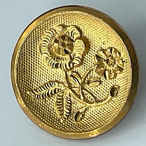 May include: A round, gold-colored button with a floral design. The button features a textured background and a raised relief of two flowers and leaves. The button is likely made of metal and has a vintage aesthetic.