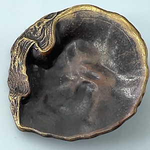 May include: A bronze-colored, shell-shaped dish with a gold-colored rim. The dish has an ornate, textured design on one side. The interior of the dish is a darker bronze color.