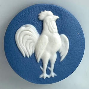 May include: A circular blue button featuring a raised white relief of a rooster in the center. The blue background has a textured, matte finish, while the white rooster figure is smooth and detailed, showing the bird in a standing profile.