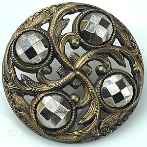 May include: A circular brass-toned button featuring an openwork design of swirling leaves and vines. Four round, faceted, metallic-colored stones are set into the metal frame, creating a reflective, checkered pattern across the surface of each stone.