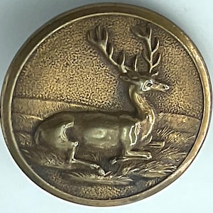 May include: A round, antique-style button with a raised relief of a deer resting in a grassy field. The button has a bronze finish and a detailed design, suitable for sewing or crafting projects.
