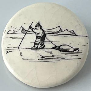 May include: A round, off-white ceramic piece with a black ink illustration of a person in winter clothing pulling a sled across a snowy landscape. The design includes mountains, a setting sun, and a signature.