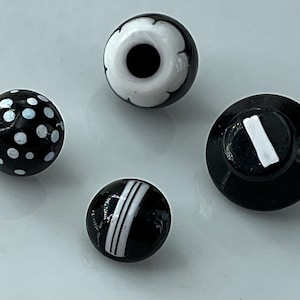May include: Four black and white decorative beads. One bead has white polka dots, another has white stripes, and a third has a white floral design. The fourth bead is a black button with a white stripe.