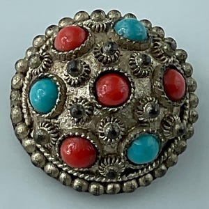 May include: A round, silver-toned button with a detailed floral design. The button features alternating red and turquoise-colored stones, with smaller dark accents. The outer edge is lined with small, silver-colored beads.