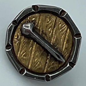May include: A circular button featuring a textured gold-toned center disc with a raised, dark metallic nail-shaped element positioned diagonally across it, all encased within a notched, silver-toned metallic outer rim.