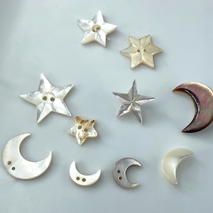May include: A collection of assorted buttons shaped like stars and crescent moons, crafted from iridescent mother-of-pearl. The buttons feature varying sizes and shades of white, cream, and dark brown, each with two small holes for attachment.