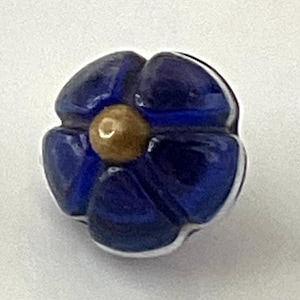 May include: A close-up of a blue flower-shaped glass bead with a golden-brown center. The bead has a glossy finish and white accents. The flower has five petals and is set against a white background.