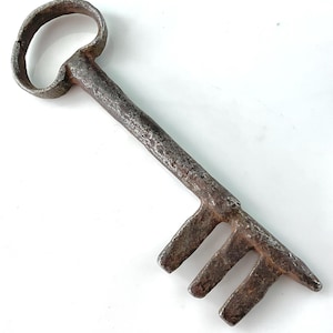 May include: A large, antique-style key made of weathered metal. The key has a rounded handle and a long stem with three teeth. The metal has a rustic, aged appearance, suggesting it is vintage or a replica.