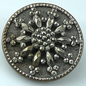 May include: A round, metallic button featuring a dark, textured background with a raised, silver-toned floral or star-shaped design in the center. The surface is embellished with faceted, reflective accents and a beaded border.