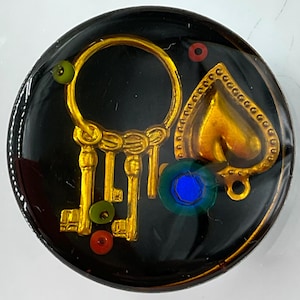 May include: A circular black resin cabochon featuring embedded gold-toned metal charms, including a ring of keys and a heart shape. The design is accented with small, colorful beads in shades of green, red, and a single prominent blue circular stone.