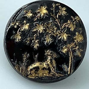 May include: A round, black button with a gold-colored relief design. The design features a deer resting under a tree with gold-colored flowers. The button has a glossy finish.