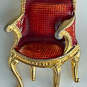 May include: A miniature gold-colored chair pin with a red seat and back. The chair has ornate detailing and curved legs, with a glossy finish. The pin is likely a decorative accessory or collectible item.