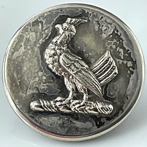 May include: A round, silver-toned button with a detailed relief of a bird perched on a branch. The bird has a crest and detailed feathers. The button has a textured, aged appearance.