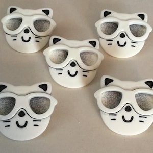 May include: Five white ceramic cat figurines with black eyes and mouths. Each cat is wearing sunglasses.